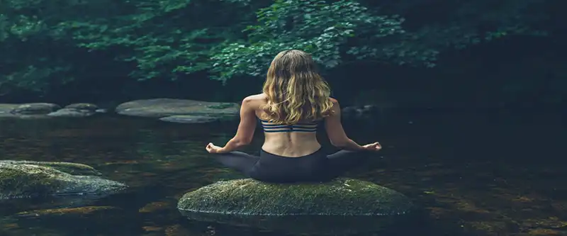 Female meditating on a rock in the middle of a river. The huge benefits of meditation and how can meditation boost our energy and vitality @ i-am-meditations.com